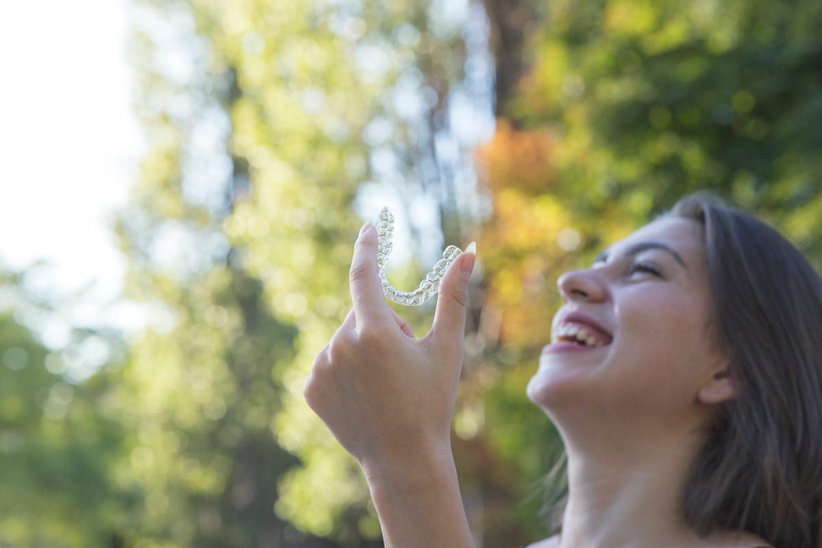 Woman smiling while holding Invisalign aligners outdoors